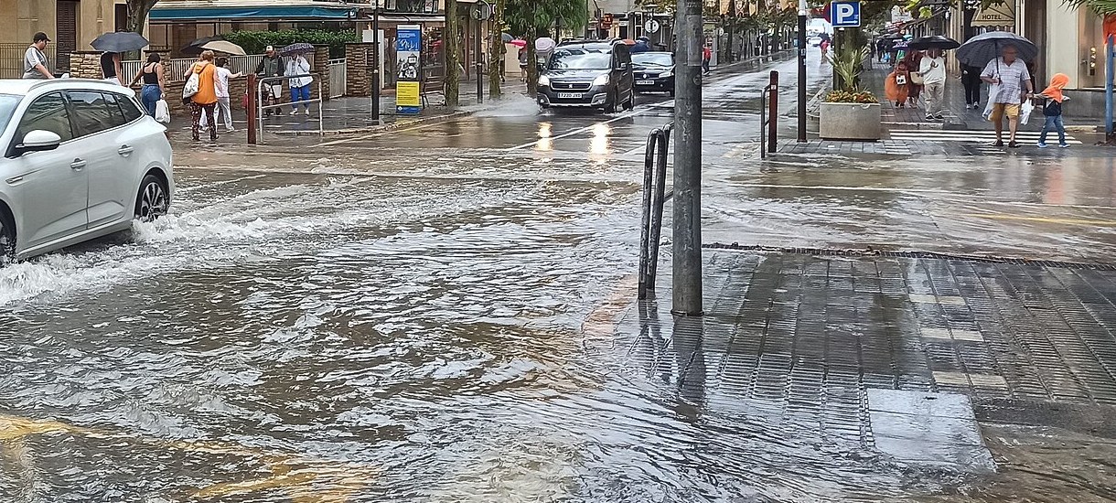 Calle de Salou inundada por una fuerte lluvia, con un coche salpicando agua y personas con paraguas al fondo