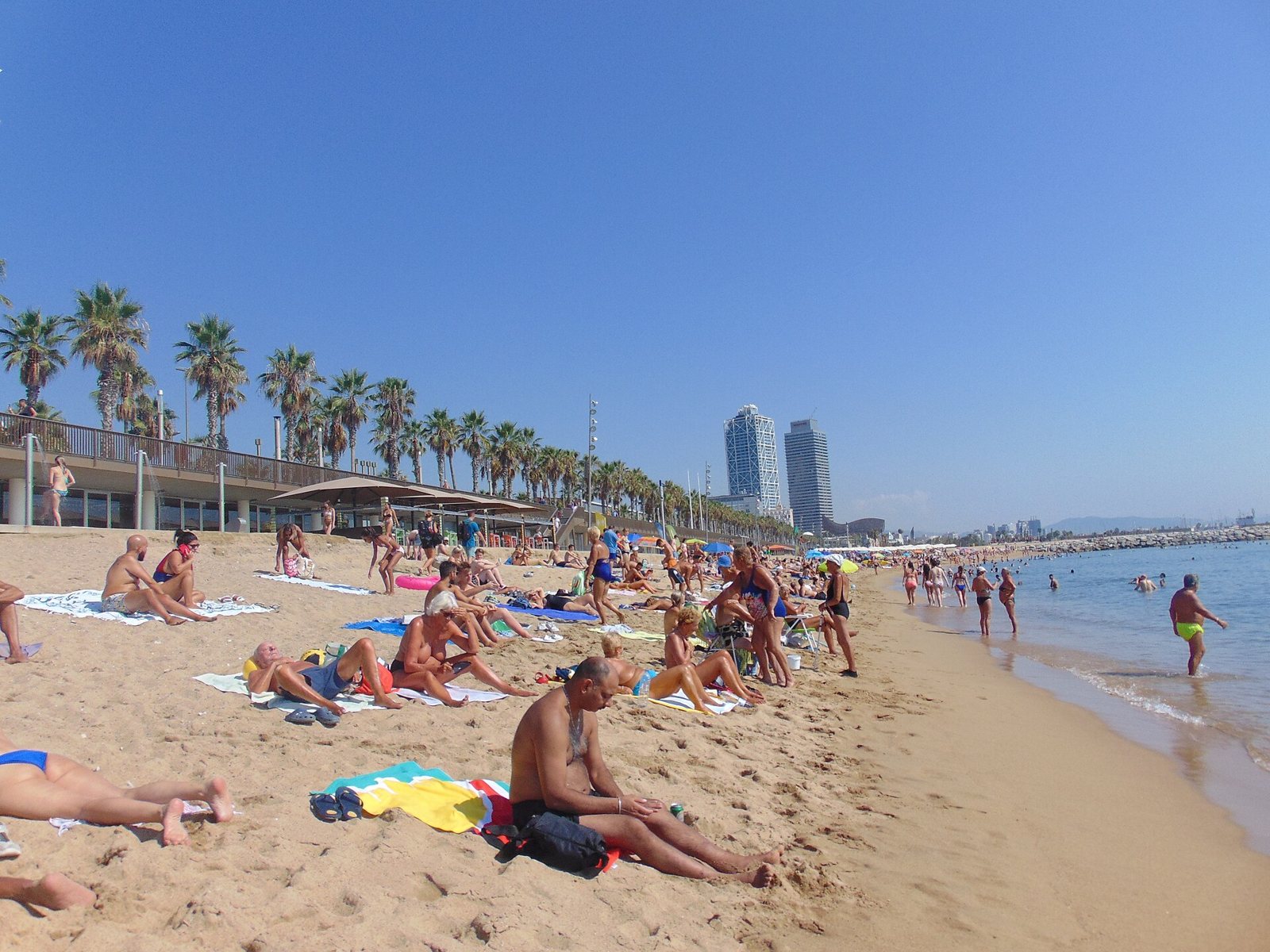 Playa de la Barceloneta llena de bañistas, con las torres del puerto olímpico al fondo
